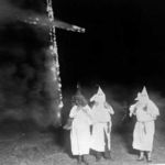 Drei Mitglieder des rassistischen Ku-Klux-Klans in ihren weißen Roben mit durch Haubenmützen verdeckten Gesichtern stehen vor einem brennenden Kreuz.