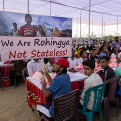 Zahlreiche Rohingya-Geflüchtete sitzen bei einer Iftar-Veranstaltung im Distrikt Cox’s Bazar in Bangladesch an langen Tischen unter einem Festzelt. Im Vordergrund hält ein Mann ein Schild mit der Aufschrift „We Are Rohingya, Not Stateless!“. An dem gemeinsamen Fastenbrechen nahmen rund 60.000 Geflüchtete und UN-Generalsekretär António Guterres teil.